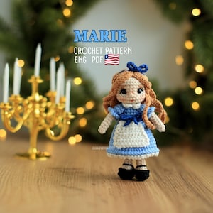 Può includere: Una bambola all'uncinetto con capelli castano chiaro, un fiocco blu e un vestito blu e bianco. Il testo MARIE CROCHET PATTERN ENG PDF con un'icona della bandiera USA è visibile. La bambola ha scarpe nere e poggia su una superficie in legno.