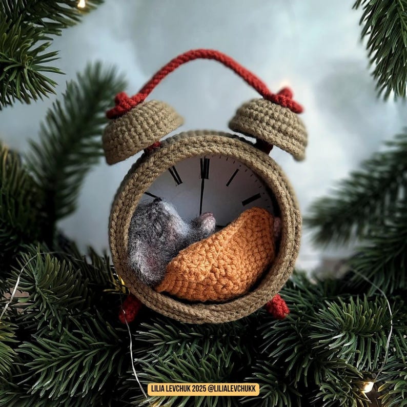 May include: A handcrafted crochet alarm clock with a sleeping mouse inside, nestled on a crocheted orange blanket. The clock is beige with red accents and sits among evergreen branches. The text "LILIA LEVCHUK 2025 @LILIALEVCHUKK" is visible.