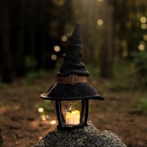 May include: A black crochet witch hat lantern with a brown belt and buckle. The lantern has a glass enclosure with a lit candle inside. The lantern is sitting on a rock in a forest setting.