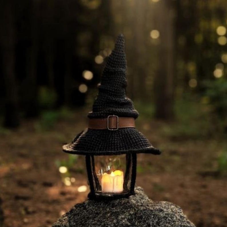 May include: A black crochet witch hat lantern with a brown belt and buckle. The lantern has a glass enclosure with a lit candle inside. The lantern is sitting on a rock in a forest setting.