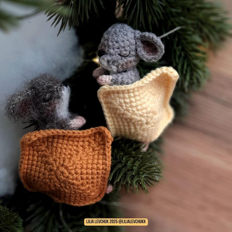 May include: Two crocheted gray mice, each nestled in a small, open-topped pouch. One pouch is a light yellow, the other is a warm orange. The mice are positioned on a bed of green pine needles, creating a cozy, handmade aesthetic.