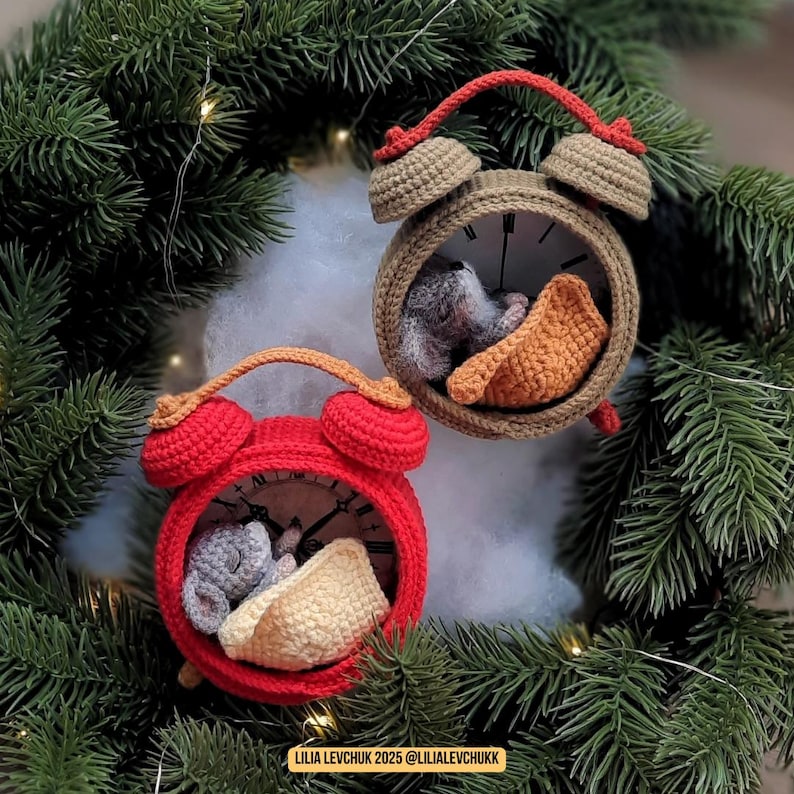 May include: Two crochet alarm clock ornaments, one red and one tan, each with a sleeping mouse inside. The clocks are surrounded by evergreen branches and white fluff. The red clock has a tan handle, and the tan clock has a red handle.