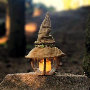 May include: A handmade, beige crochet lantern, shaped like a witch's hat. The lantern has a glass globe with a lit candle inside, emitting a warm light. The hat is decorated with a braided band and green leaves, placed on a stone.