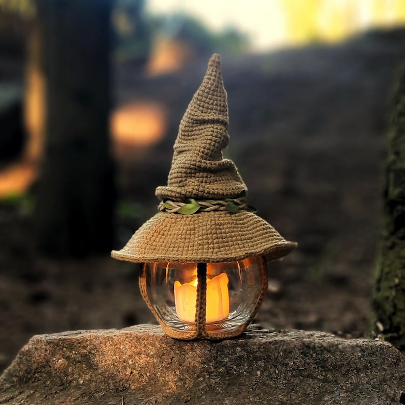 May include: A handmade, beige crochet lantern, shaped like a witch's hat. The lantern has a glass globe with a lit candle inside, emitting a warm light. The hat is decorated with a braided band and green leaves, placed on a stone.