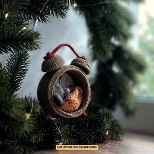 May include: A handmade crochet alarm clock in olive green, with a red handle and accents. Inside the clock face are two small, crocheted figures in gray and orange. The clock is set against a backdrop of evergreen branches and a window.