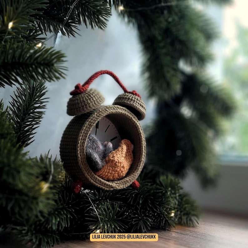 May include: A handmade crochet alarm clock in olive green, with a red handle and accents. Inside the clock face are two small, crocheted figures in gray and orange. The clock is set against a backdrop of evergreen branches and a window.