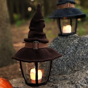 May include: A brown crochet lantern in the shape of a witch's hat, featuring a leather strap and buckle. The lantern has a glass enclosure with a lit candle inside. Another similar lantern is in the background, creating a Halloween theme.