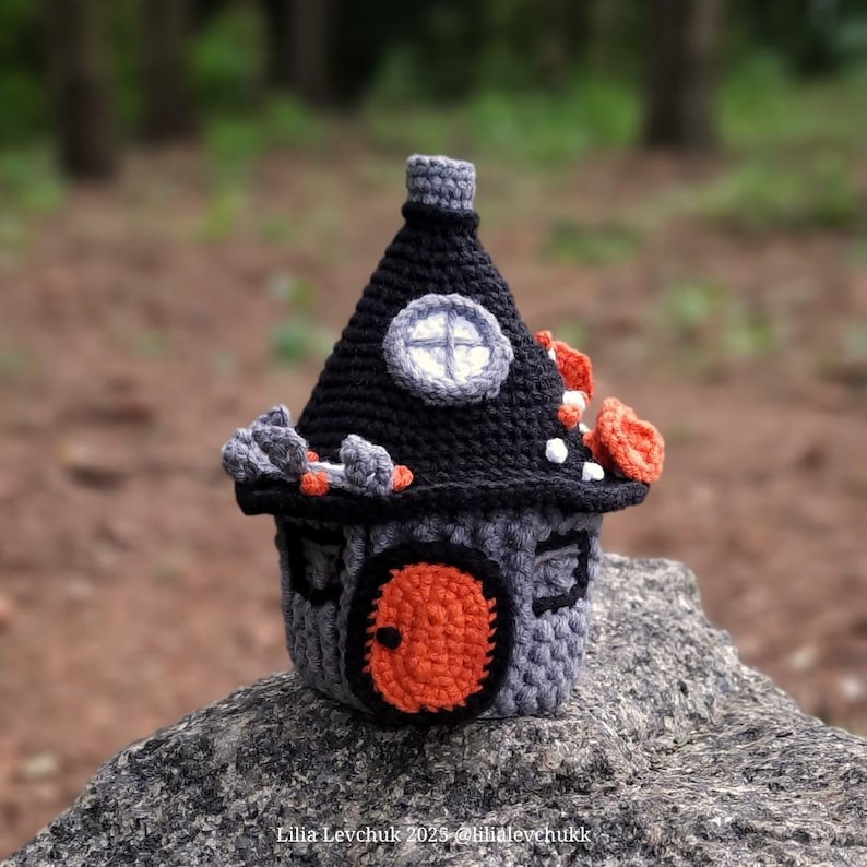 May include: A crocheted house with a black roof and gray walls, adorned with orange and gray accents. The round orange door and small window add detail to the whimsical design. The house is sitting on a rock.