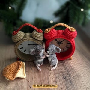 May include: Two crochet alarm clocks, one tan and one red, flank two small gray mice. The clocks have a vintage design with a bell on top. A small crochet piece of bread is on the left. The background is blurred.