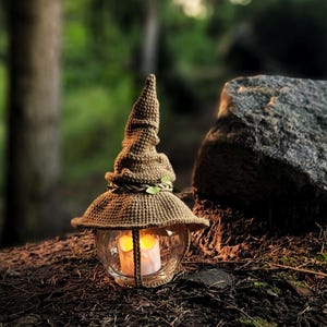 May include: A decorative lantern with a glass globe and a woven brown hat-shaped top. The lantern has a candle inside, and is decorated with a green leaf garland. The lantern is sitting on a bed of leaves and twigs.