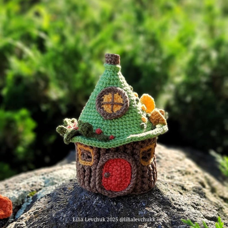 May include: A handcrafted crochet fairy house with a green roof, brown walls, and a red door. The house features small windows and decorative elements, set against a blurred green background. The text "Lilia Levchuk 2025 @lilialevchuk" is visible.