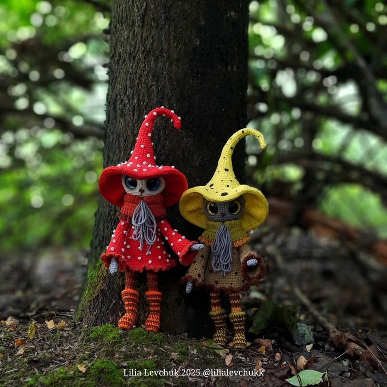May include: Two handmade crochet dolls stand near a tree. One doll wears a red hat with white polka dots, a matching scarf, and orange boots. The other doll has a yellow hat and a brown outfit. The dolls have cat-like faces.
