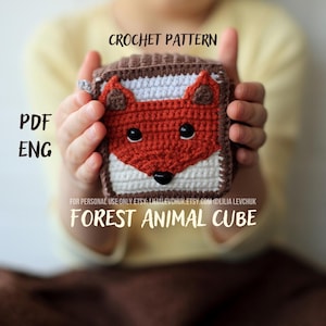 May include: A crocheted cube featuring a fox face design in shades of red, white, and brown. The cube is held in a person's hands. The text "CROCHET PATTERN", "PDF ENG", and "FOREST ANIMAL CUBE" are visible.