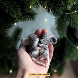 May include: Two small, handmade gray mice are nestled in a person's hand. One mouse is a darker gray with a fuzzy texture, while the other is a lighter gray with a crocheted appearance. They are embracing, with closed eyes.