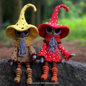 May include: Two handmade crochet dolls, one in yellow with black spots and the other in red with white spots. Both dolls have large eyes, hats, scarves, and detailed clothing. The dolls are sitting on a rock.