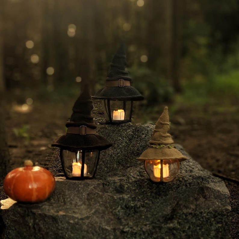 May include: Three glass lanterns with lit candles, each topped with a witch's hat, are arranged on a large rock. A small orange pumpkin is also present. The lanterns are black with brown hatbands, perfect for Halloween.
