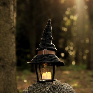 May include: A black crocheted witch hat lantern with a brown leather strap and buckle. The lantern has a glass enclosure with a lit candle inside, resting on a stone in a forest setting. The hat's pointed top and wide brim create a whimsical, decorative piece.