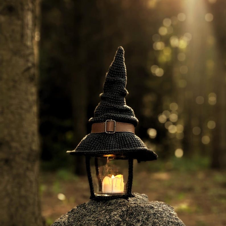 May include: A black crocheted witch hat lantern with a brown leather strap and buckle. The lantern has a glass enclosure with a lit candle inside, resting on a stone in a forest setting. The hat's pointed top and wide brim create a whimsical, decorative piece.