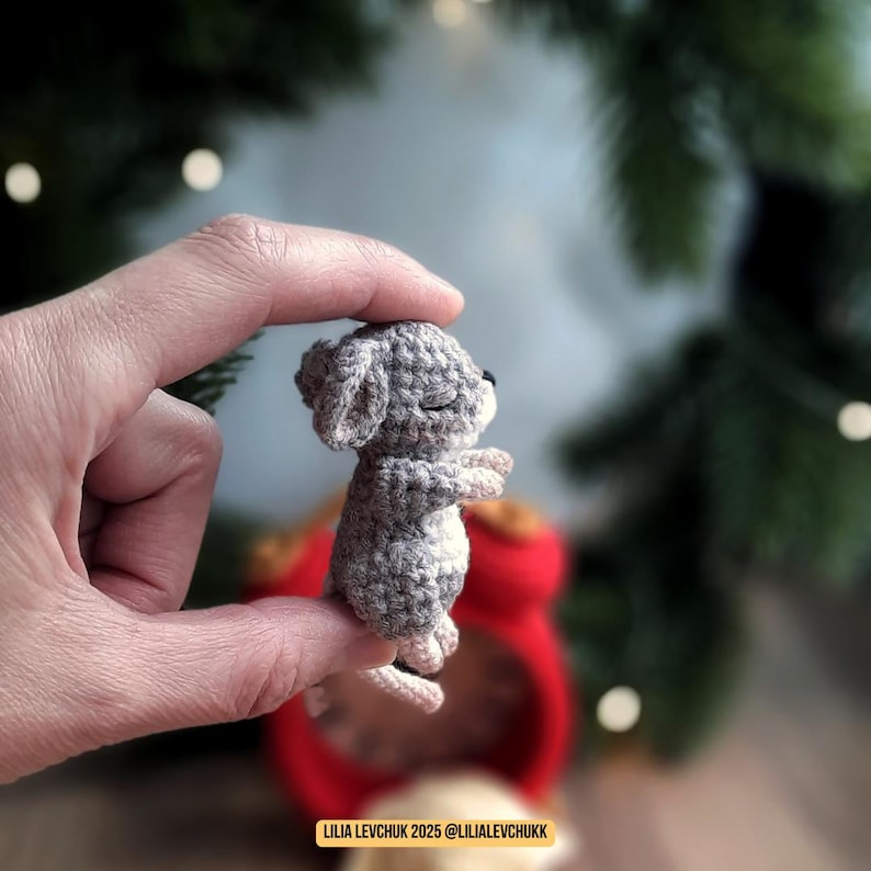 May include: A small, crocheted gray mouse figurine with closed eyes and outstretched arms. The mouse has a white belly and pink ears and feet. The figurine is held between fingers, with a blurred red object in the background. The text "LILIA LEVCHUK 2025 @LILIALEVCHUKK" is at the bottom.