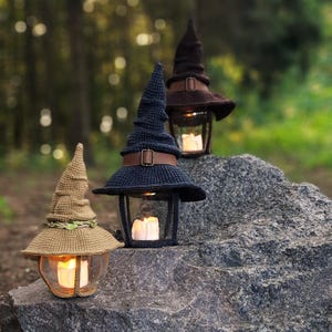 May include: Three decorative lanterns, each topped with a crocheted witch hat in brown and navy blue. The hats feature leather straps. Inside the glass enclosures, lit candles provide a warm light. The lanterns are displayed on a large, gray rock, creating a whimsical scene.