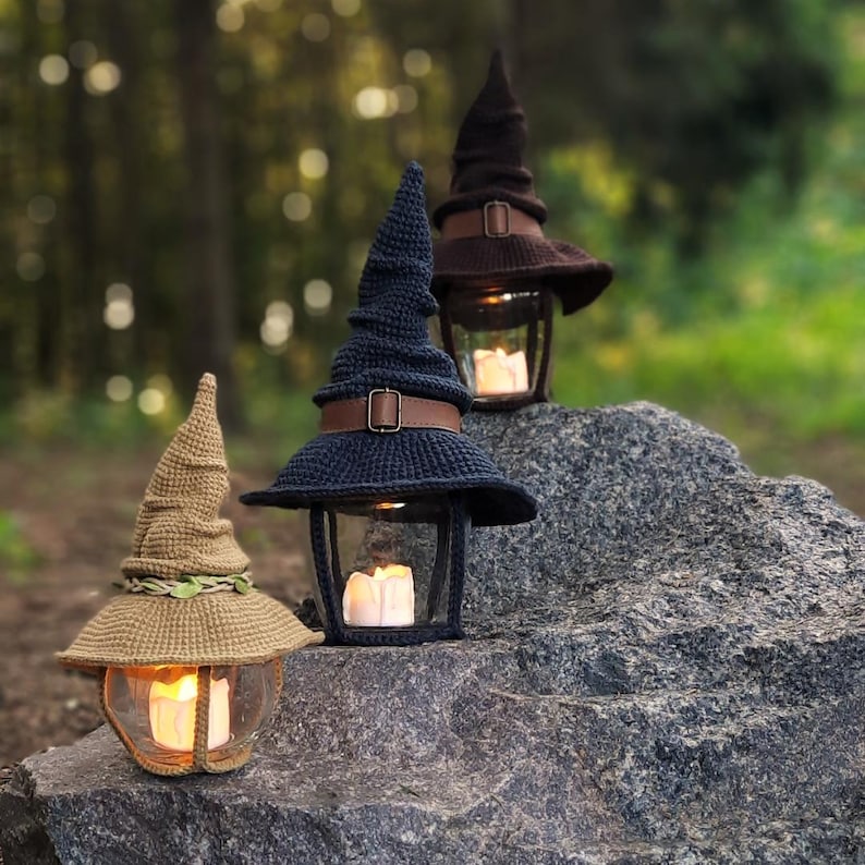 May include: Three decorative lanterns, each topped with a crocheted witch hat in brown and navy blue. The hats feature leather straps. Inside the glass enclosures, lit candles provide a warm light. The lanterns are displayed on a large, gray rock, creating a whimsical scene.