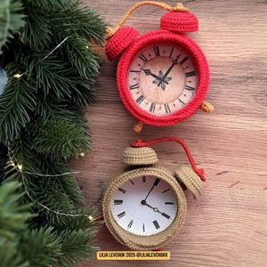 May include: Two crochet alarm clocks, one red and one tan, sit on a wooden surface. The red clock has a vintage-style face with Roman numerals. The tan clock has a white face with black hands. Both clocks have bells and handles.