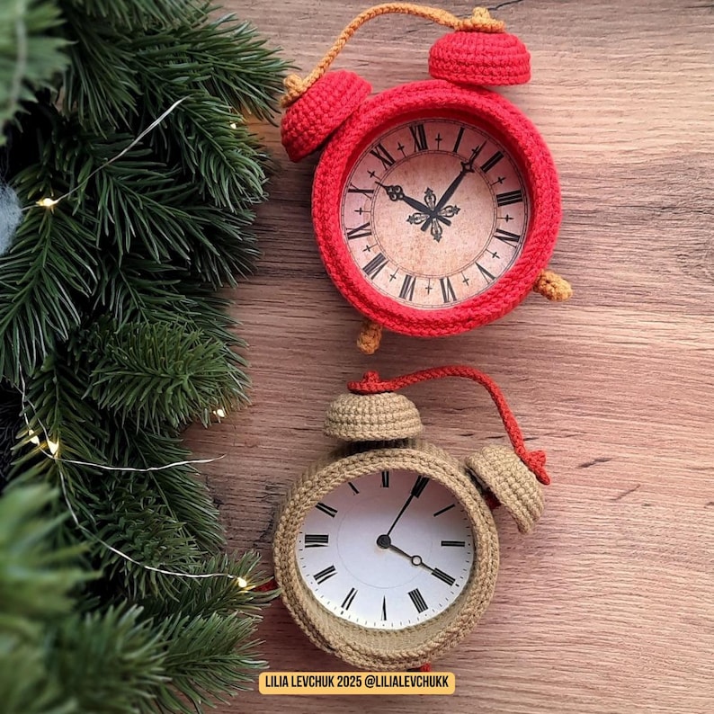 May include: Two crochet alarm clocks, one red and one tan, sit on a wooden surface. The red clock has a vintage-style face with Roman numerals. The tan clock has a white face with black hands. Both clocks have bells and handles.