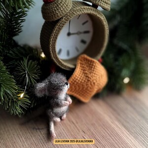 May include: A handmade crochet alarm clock in olive green with a white clock face, a small gray mouse figurine, and an orange crochet mask. The scene is set on a wooden surface with a backdrop of green pine branches.