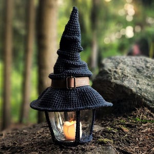 May include: A navy blue crocheted lantern with a witch hat design. The lantern has a glass enclosure with a lit candle inside. A brown leather belt with a buckle encircles the lantern. The lantern is sitting on a bed of leaves and dirt.