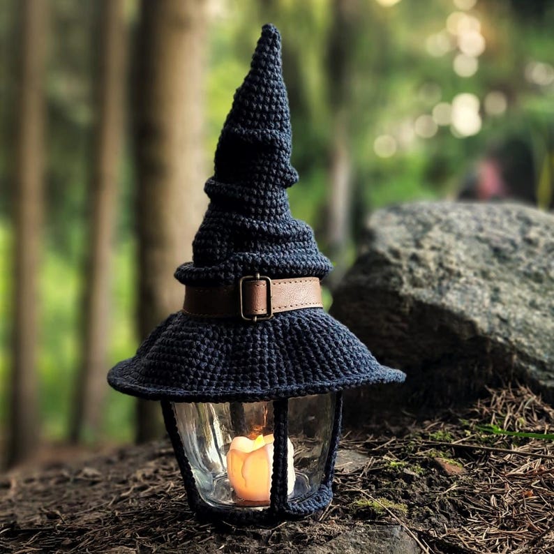 May include: A navy blue crocheted lantern with a witch hat design. The lantern has a glass enclosure with a lit candle inside. A brown leather belt with a buckle encircles the lantern. The lantern is sitting on a bed of leaves and dirt.