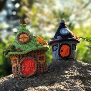 May include: Two crocheted fairy houses. One is brown and green with a red door and a round window. The other is gray and black with an orange door and a round window. Both have decorative details and sit on a rock.