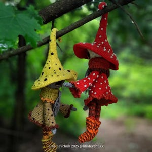 May include: Two handmade crochet mushroom figures hanging from a tree branch. One figure wears a yellow hat with black spots and a brown outfit. The other figure is dressed in a red mushroom cap with white spots and a matching red outfit. The background is blurred green foliage.