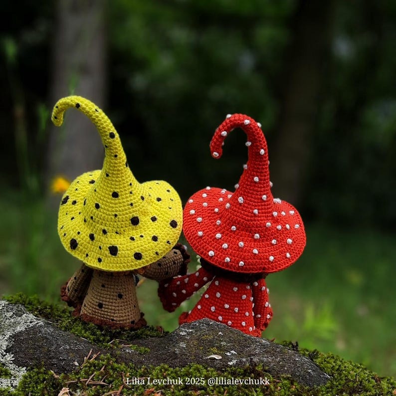 May include: Two handmade crochet mushroom figures. One mushroom has a yellow cap with black spots, the other has a red cap with white spots. Both have brown stems and are set on a mossy rock.