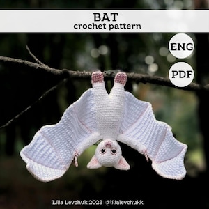 May include: A white crochet bat hangs upside down from a tree branch. The bat has pink accents on its ears and feet, with detailed wings. The image includes the text "BAT crochet pattern" and "ENG PDF".