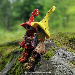 May include: Two handmade crochet dolls with whimsical witch-like features. One doll wears a red hat with white polka dots, the other a yellow hat with black dots. Both dolls have long, pointed hats, and are seated on a moss-covered rock.