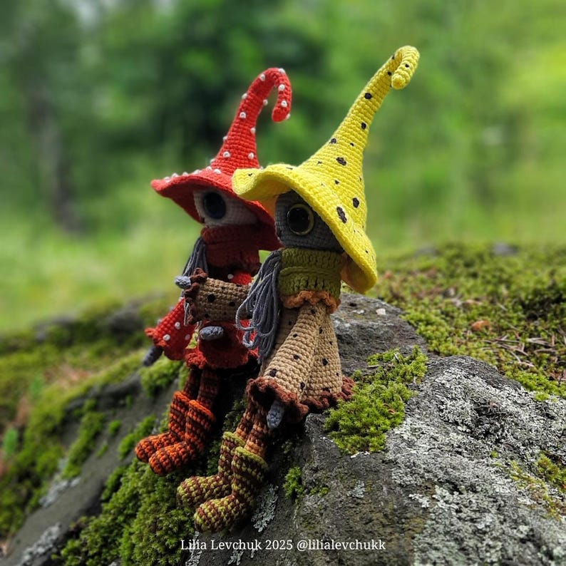 May include: Two handmade crochet dolls with whimsical witch-like features. One doll wears a red hat with white polka dots, the other a yellow hat with black dots. Both dolls have long, pointed hats, and are seated on a moss-covered rock.