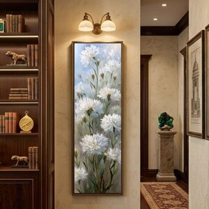 May include: A long, framed painting of white flowers with green stems and leaves, set against a pale blue and green backdrop. The artwork hangs in a hallway with a warm colour scheme, a bookcase, and a decorative light above.