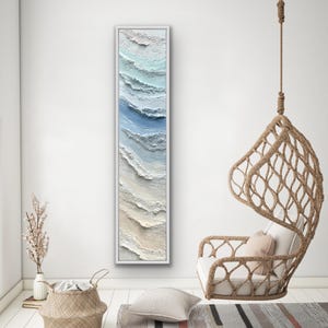 May include: A long, vertical framed artwork with a textured, abstract depiction of ocean waves. The artwork features shades of blue, white, and beige, creating a sense of depth and movement. The frame is white, and the artwork is displayed in a bright, minimalist interior.
