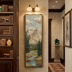 May include: A framed landscape painting depicts a mountain range, river, and evergreen trees. The artwork uses textured brushstrokes in shades of beige, green, and blue. The painting is displayed in a hallway with a bookshelf and decorative lighting.