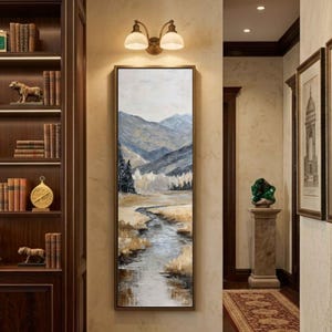 May include: A framed landscape painting depicts a serene mountain scene. The artwork features a river winding through a valley, with mountains in the background. The color palette includes shades of gray, blue, and brown, creating a calming atmosphere. The painting is displayed in a hallway.