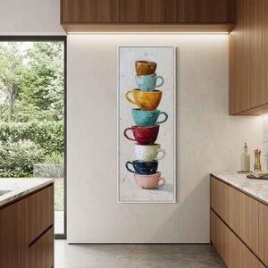 May include: A vertical rectangular wall art piece featuring a stack of seven ceramic coffee cups in various colors including brown, light blue, yellow, teal, red, white, and dark blue, resting on a light pink cup at the base.
