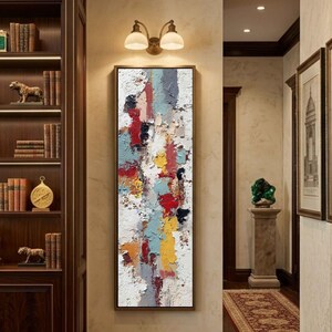 May include: Abstract art painting with a textured surface. The artwork features vertical strokes of red, yellow, blue, and white. The painting is framed in a dark brown frame and hangs on a beige wall. The artwork is displayed in a hallway.