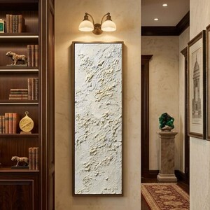 May include: A rectangular abstract textured painting in a dark brown frame. The artwork features a white base with raised, textured areas in shades of beige and cream. The painting is displayed in a hallway with a bookshelf and a decorative light fixture.