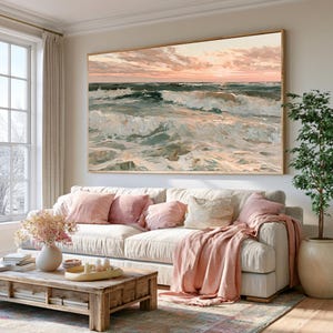 May include: A large rectangular wall art print featuring an oil painting of a turbulent ocean scene at sunset with soft pink clouds above dark teal waves, displayed in a thin wooden frame above a beige sofa with pink and cream throw pillows.
