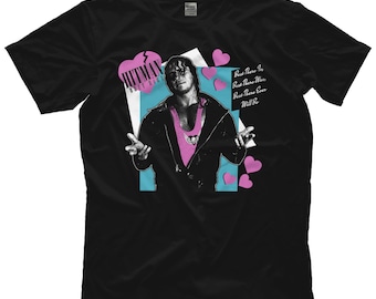 May include: Black t-shirt with a graphic of a wrestler in a pink and black wrestling outfit. The graphic is surrounded by pink hearts and the text "Hitman Best Hair" and "Best There Is, Best There Was, Best There Ever Will Be".