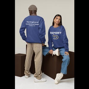 May include: Two people wearing blue sweatshirts. One sweatshirt has the text "The kingdom of heaven is at hand. Matthew 4:17" on the back. The other sweatshirt has the word "REPENT" with a curved arrow. The person on the left is wearing tan pants and the person on the right is wearing blue jeans.