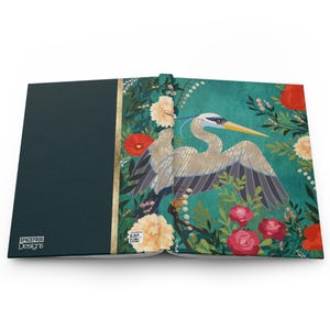 May include: Open journal with a teal cover and a gold spine. The right page features a heron illustration surrounded by flowers in shades of red, cream, and green. The left page is solid teal with the text "SPACEFROG Designs".