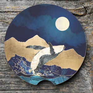 May include: Round car coaster featuring a whale breaching the water under a full moon. The design includes gold foil accents, blue water, and a dark blue night sky. The coaster is designed to fit in a car cup holder.