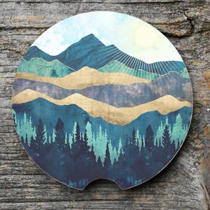 May include: A round car coaster featuring a mountain landscape design. The coaster has a blue, teal, and gold color scheme with a forest at the bottom and a sunburst in the sky. The coaster is made of absorbent material.