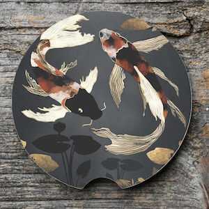 May include: A black car coaster featuring two koi fish in a circular design. The fish are depicted in shades of black, orange, and white, with gold accents. The coaster has a dark gray background with gold leaf details.